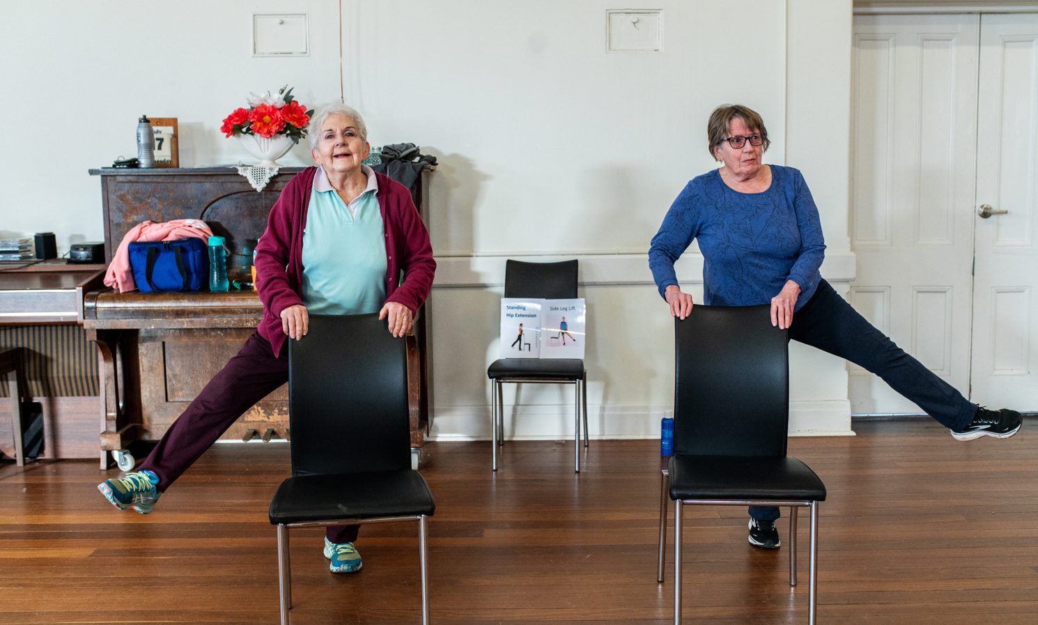 Two older women in a hall balancing on one leg with the other leg in the air while holding onto chairs as part of an exercise program.